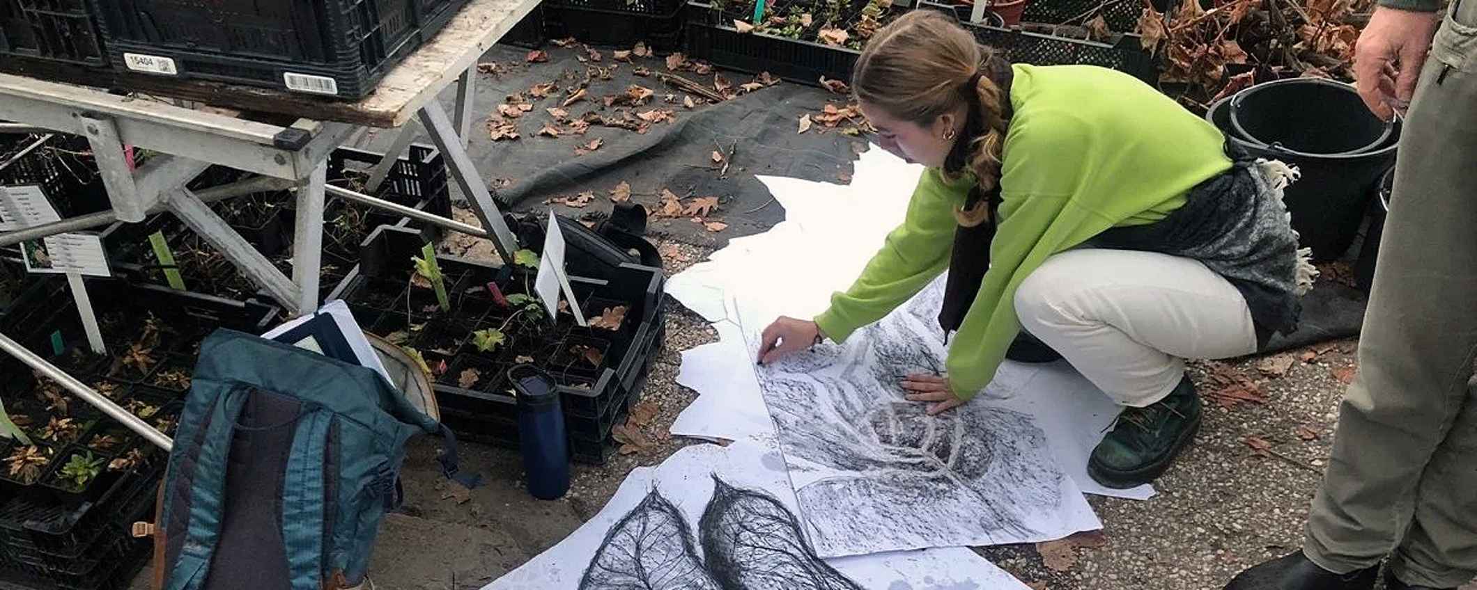 Een student zit op haar hurken buiten en tekent op grote vellen papier.